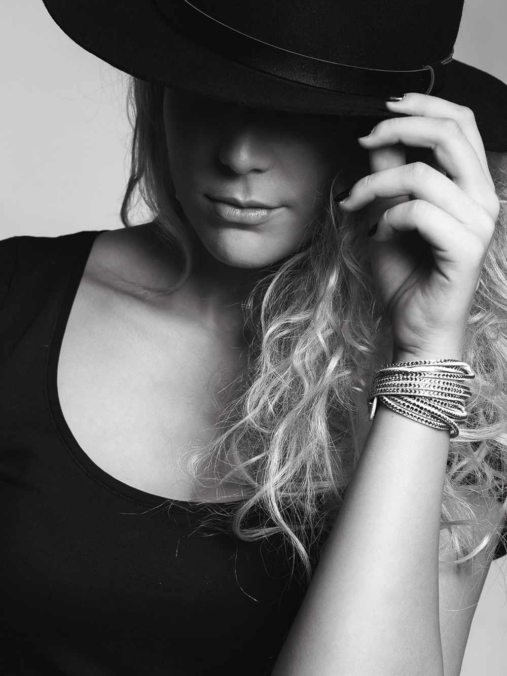 Black and white portrait of blonde woman in black top adjusting wide brim hat with bracelets.