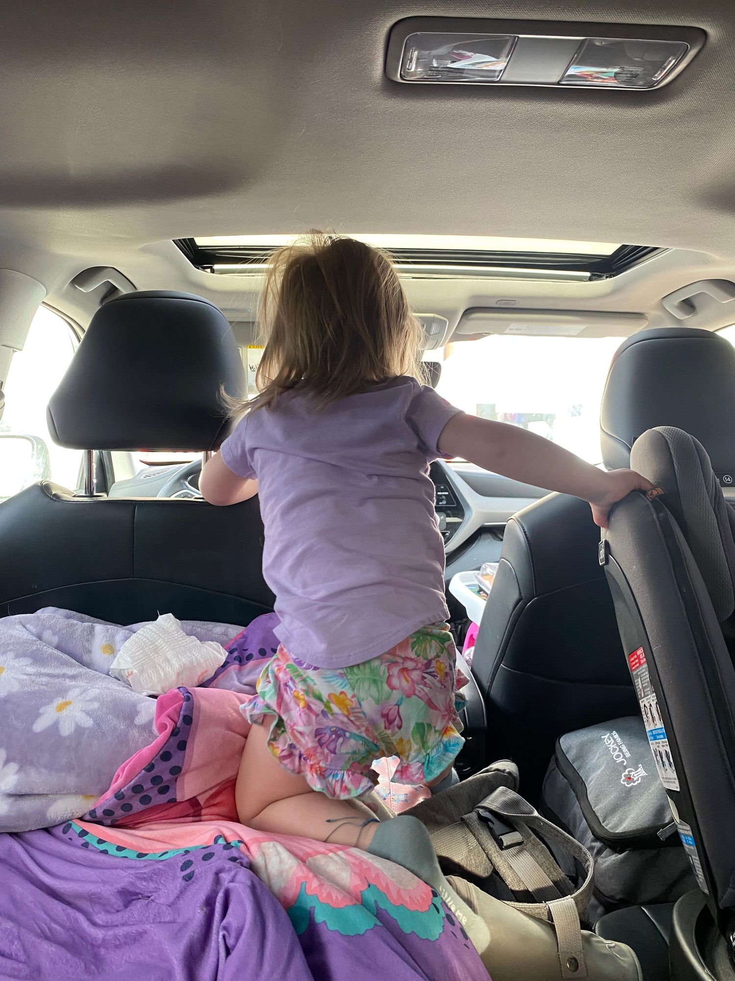Young girl in purple shirt and floral skirt standing in back seat of SUV with sunroof and purple bedding.