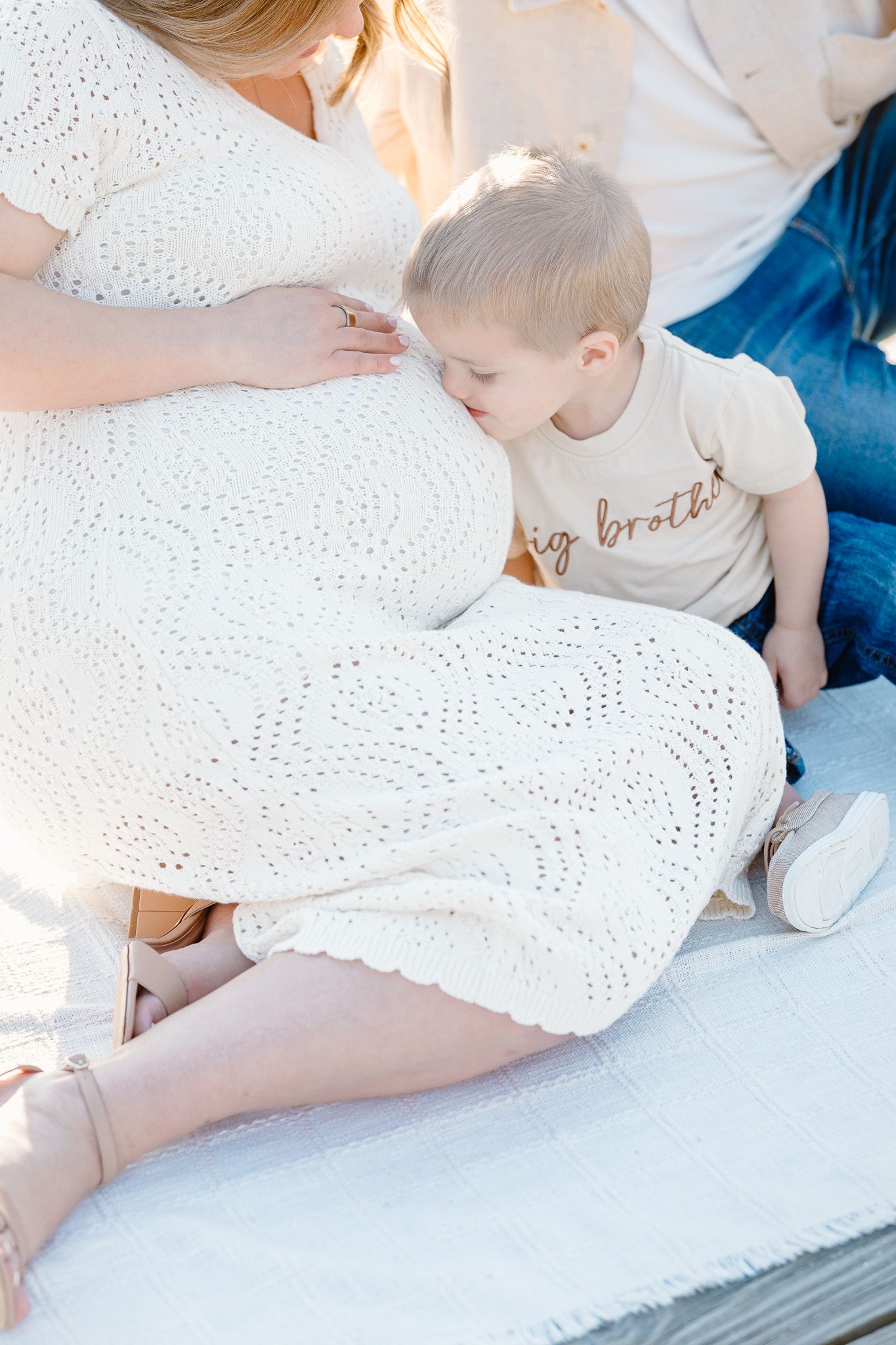 Pregnant woman in white lace dress stands on wooden boardwalk as man lifts toddler in sunny outdoor setting.