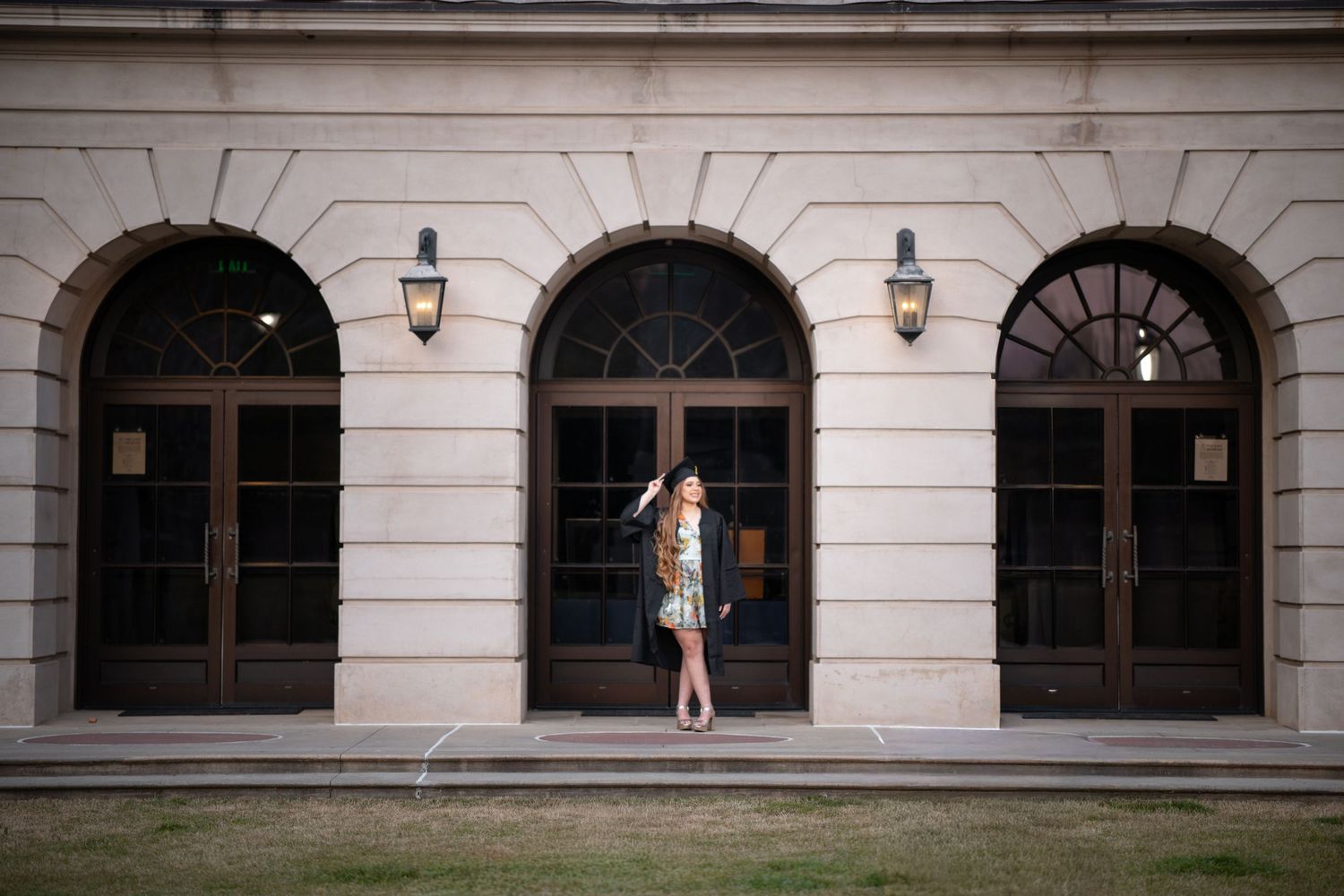 Young woman in floral dress and black hat poses before an elegant arched doorway with vintage lanterns.