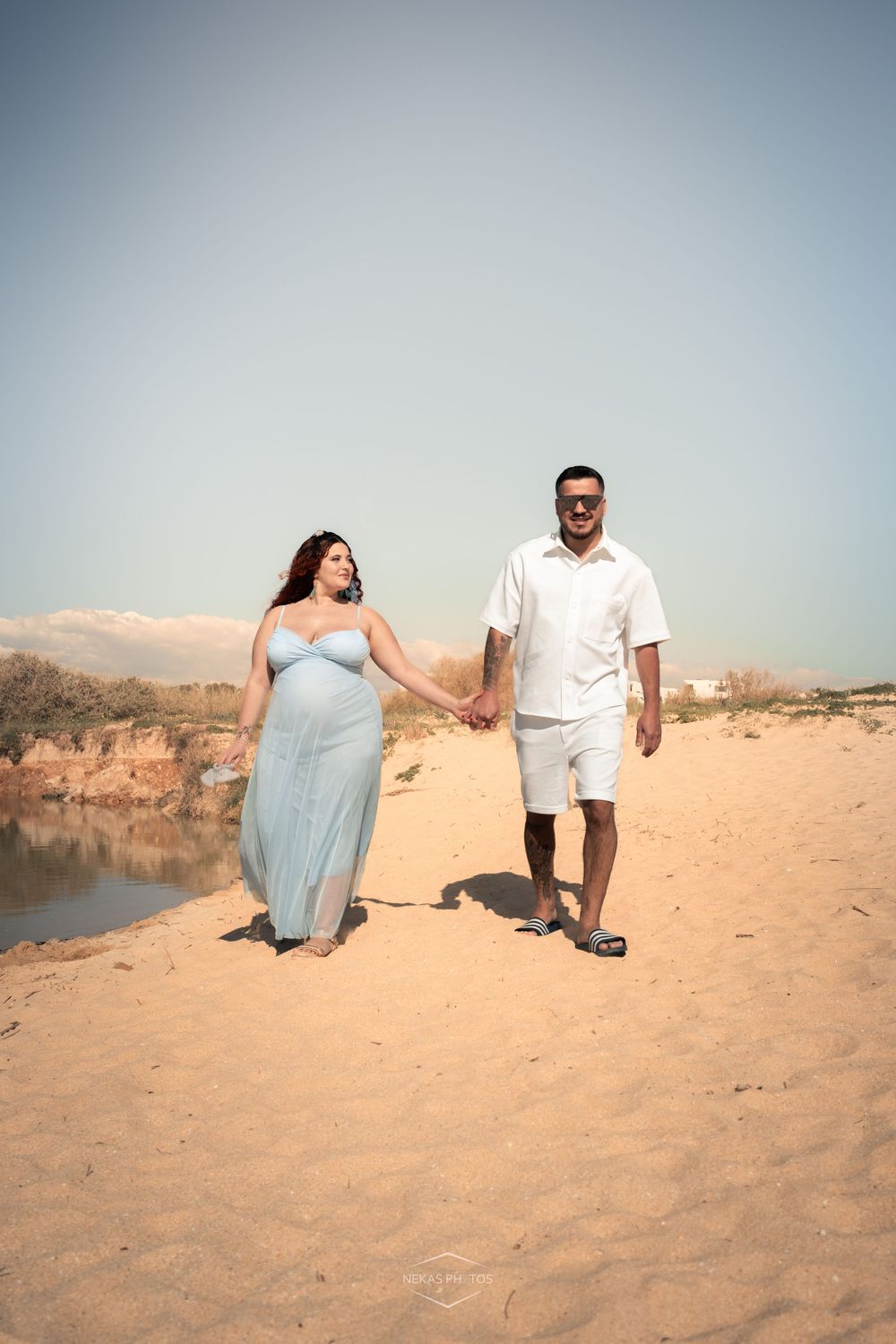 Pregnant woman in blue dress holds hands with man in white outfit while walking on sandy beach.