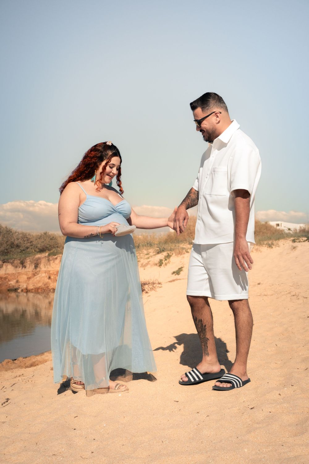 Pregnant woman in blue dress holds hands with man in white outfit on sandy beach near water.
