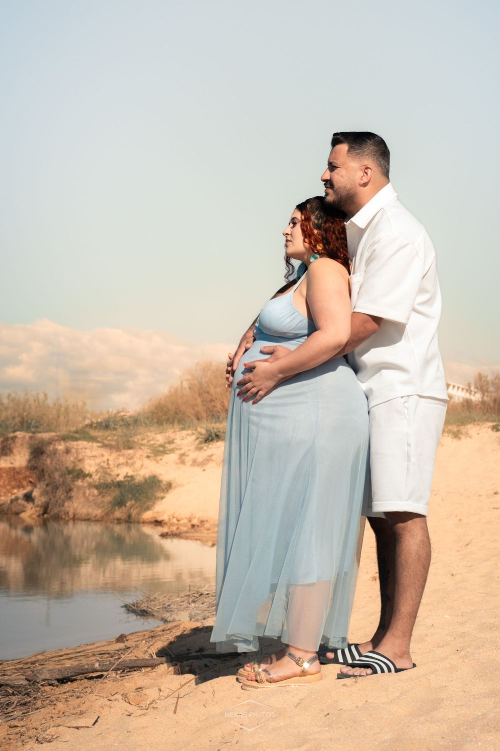Pregnant couple embracing on sandy beach, woman in blue dress, man in white outfit, serene water backdrop.