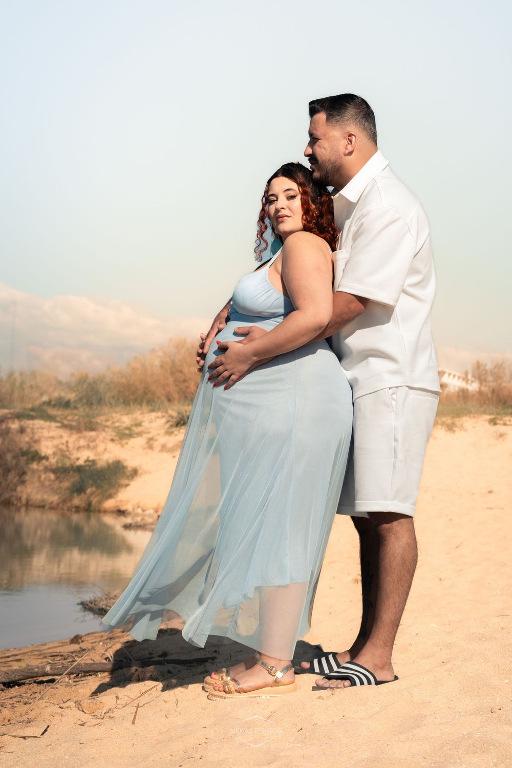 Pregnant woman in blue dress embraced by partner on sandy beach near water at sunset.