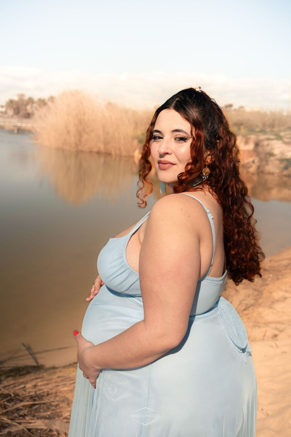Pregnant woman in light blue dress holding belly by a serene river, with long curly hair and elegant earrings.