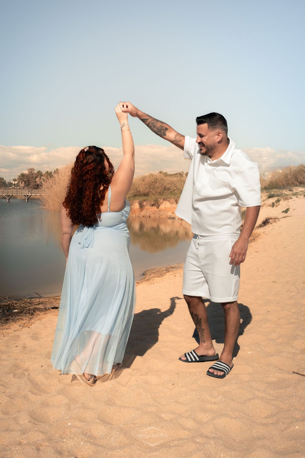 Couple dancing on sandy beach, woman in blue dress, man in white outfit, with calm lake and dunes in background.