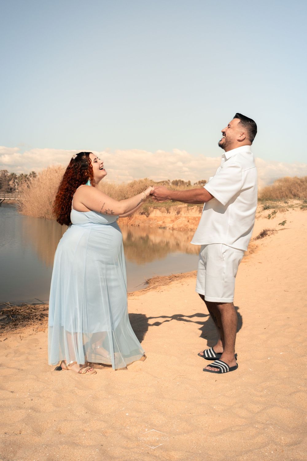 A joyful couple holds hands on a sandy beach, woman in light blue dress, man in white outfit, with water in background.
