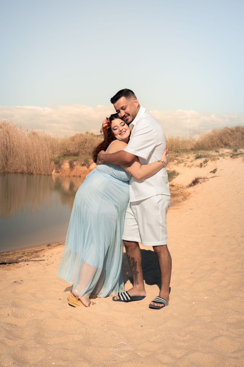 Couple embracing tenderly on sandy beach, pregnant woman in blue dress, man in white, near serene lake.