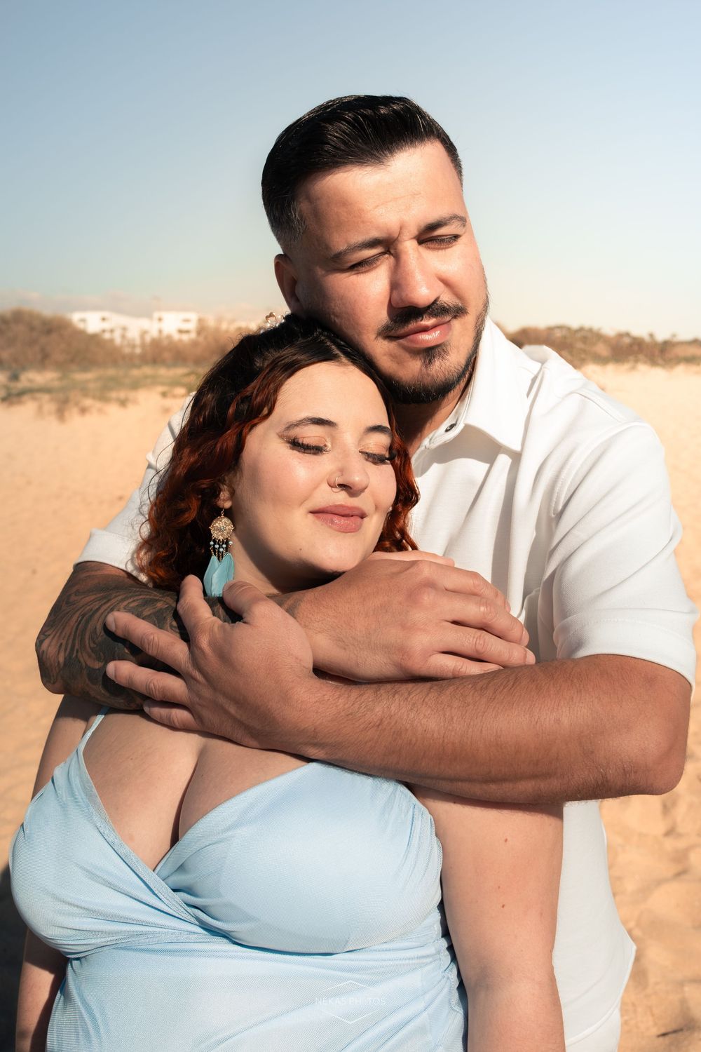 A couple embracing on a sandy beach, woman in light blue dress with red hair, man in white shirt with tattoos.