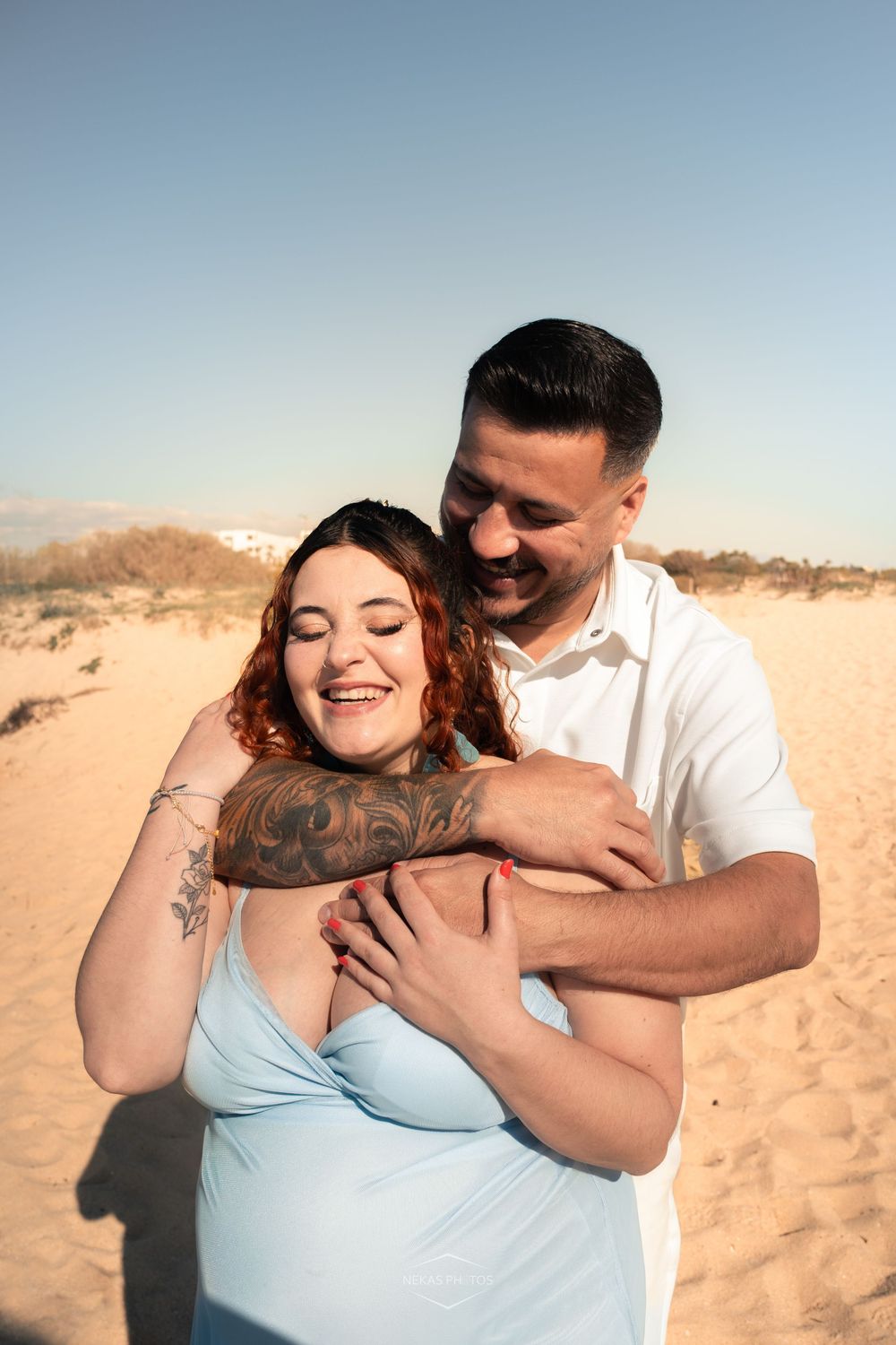 A smiling couple embraces on a sandy beach, woman with tattoos in blue dress, man in white polo at golden hour.