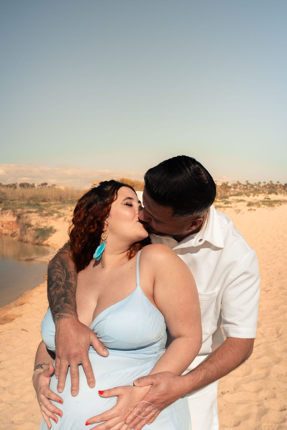 Pregnant couple sharing a kiss on a sandy beach, woman in blue dress with tattoos, man in white shirt cradling her baby bump.