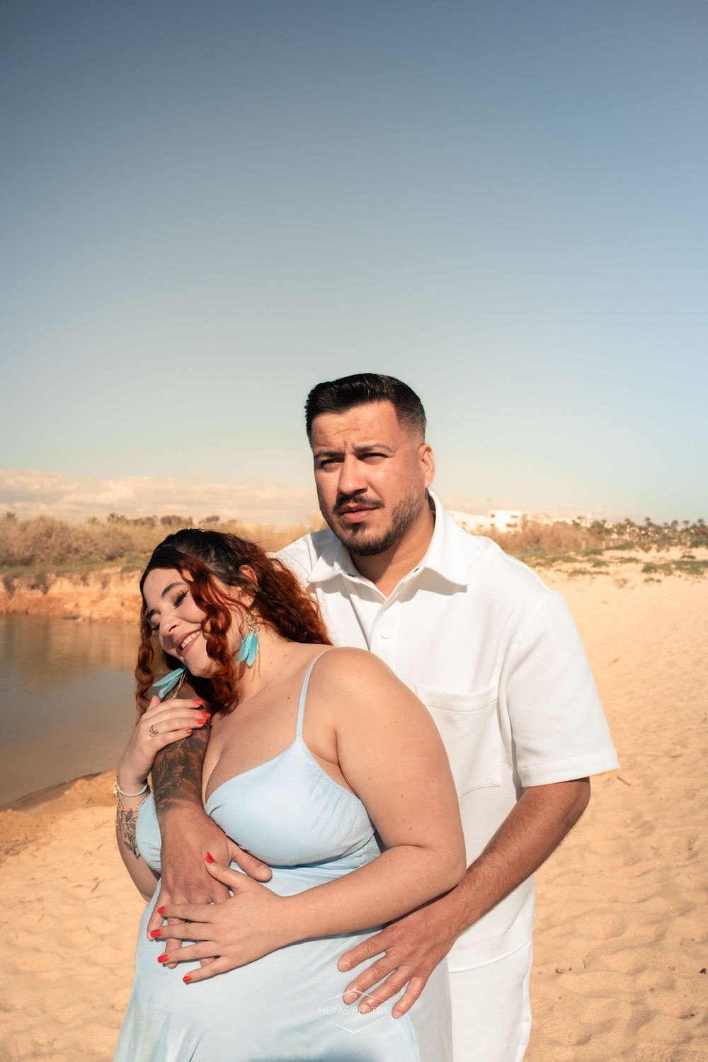 A couple embraces on a sandy beach, the woman in a blue dress smiling, the man in white holding her from behind.