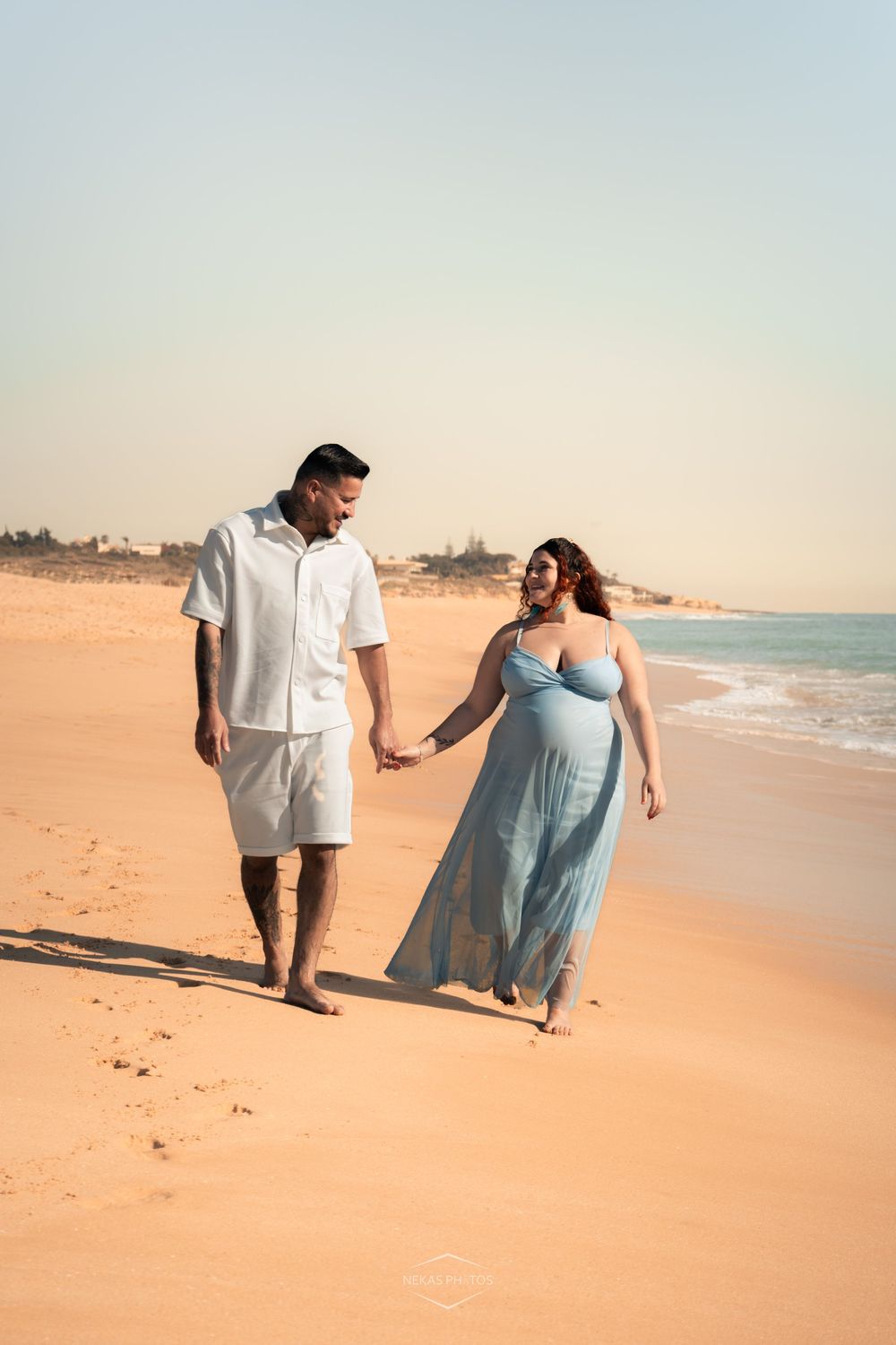 Pregnant couple holding hands walking on a sandy beach, woman in light blue maxi dress, man in white outfit.