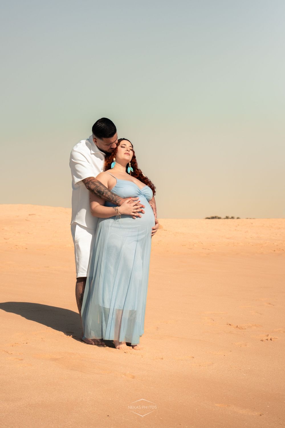 Pregnant woman in light blue dress embraced by partner in white on sandy desert dunes during maternity photoshoot.
