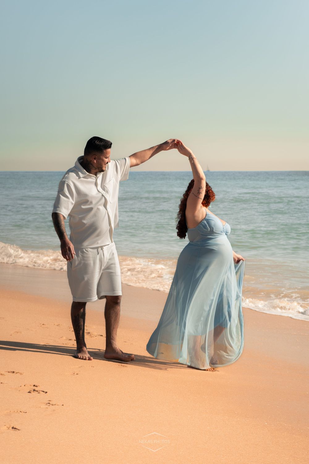 Couple dancing on a sandy beach at sunset, man in white outfit spinning woman in flowing light blue dress by the ocean.