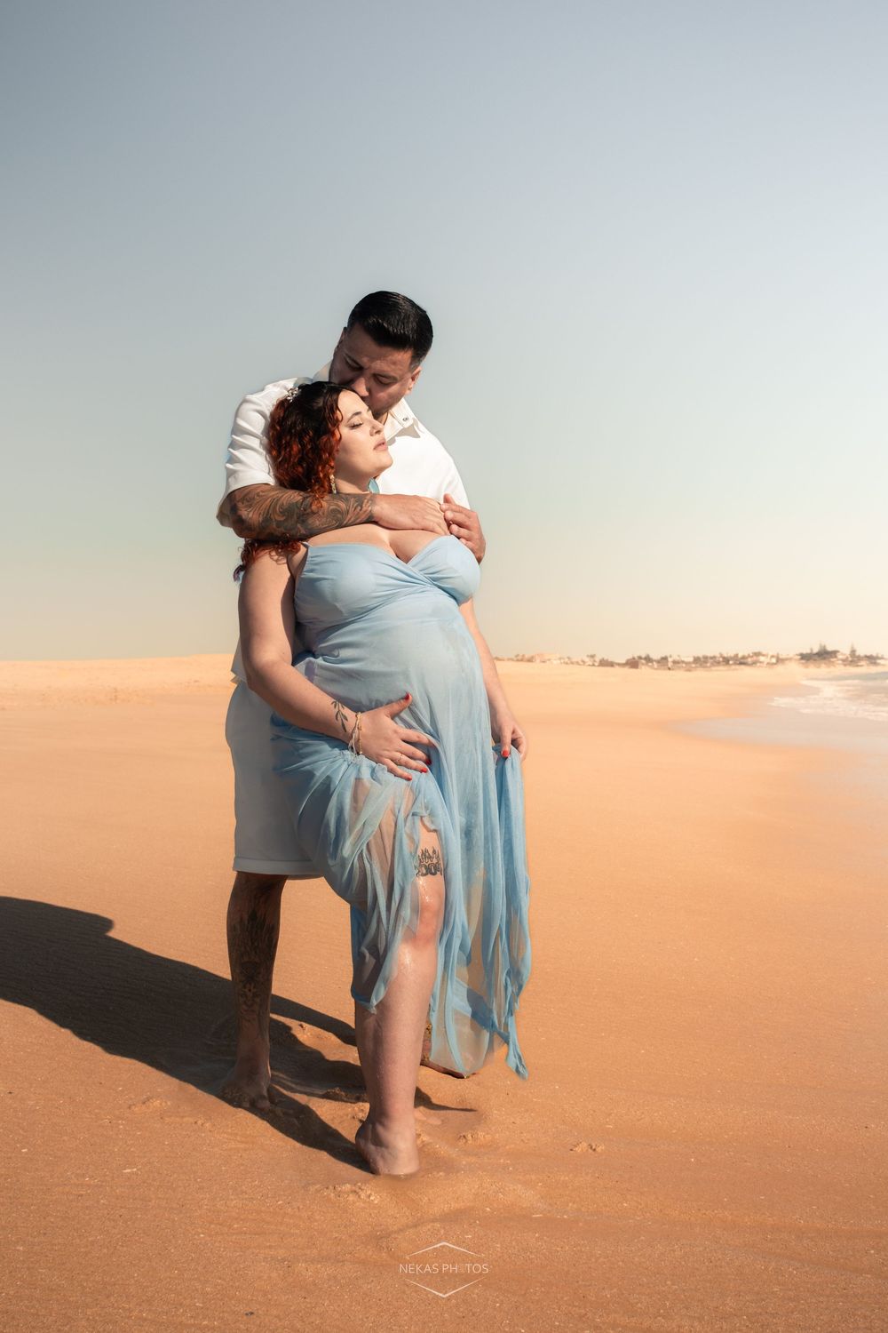 Couple embracing on sandy beach at sunset, pregnant woman in flowing blue dress, man in white shirt kissing her forehead.
