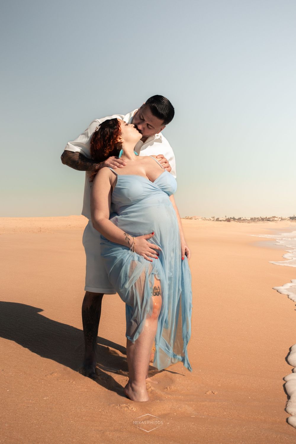 Pregnant woman in blue dress kissing partner on sandy beach at sunset.