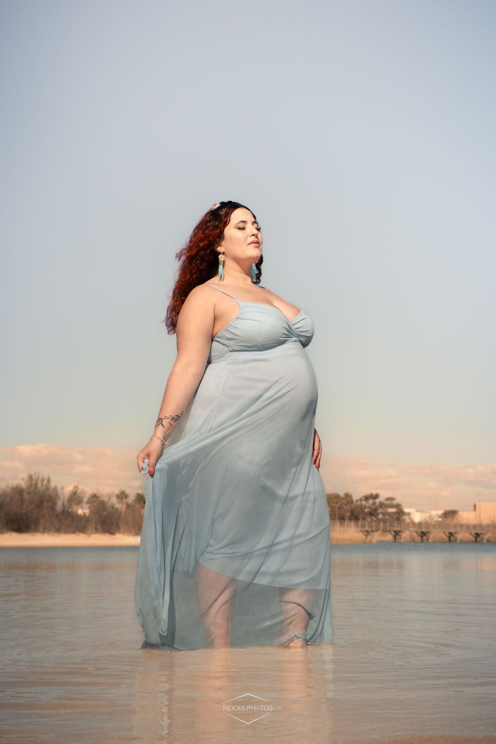 Pregnant woman in flowing light blue dress standing in shallow water during golden hour.