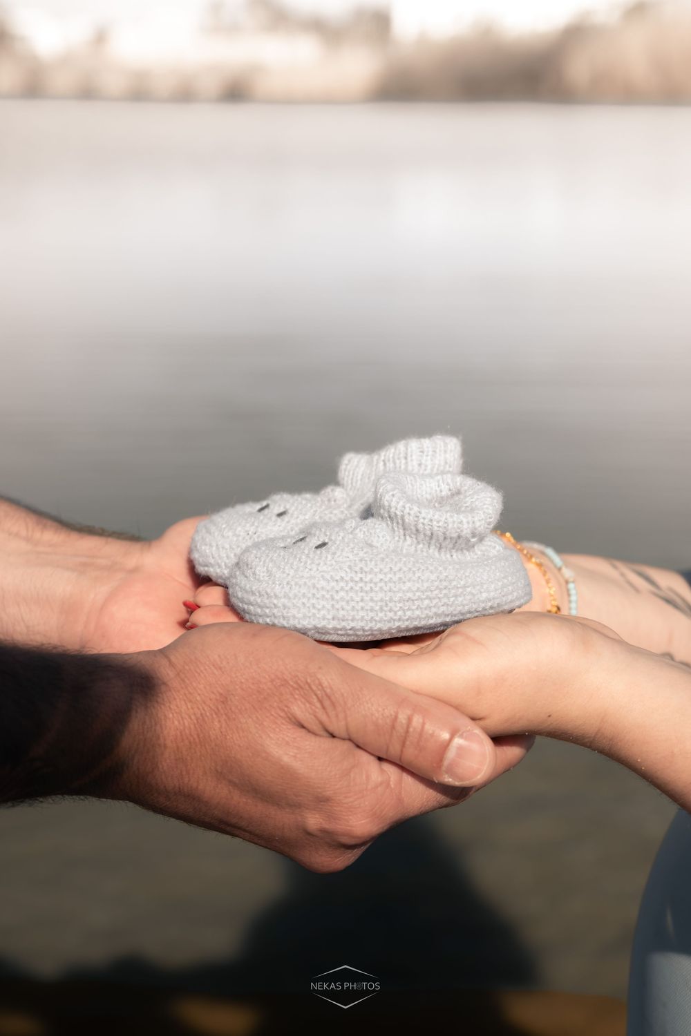 Two hands gently holding tiny gray knitted baby booties near a serene lake, announcing a pregnancy.