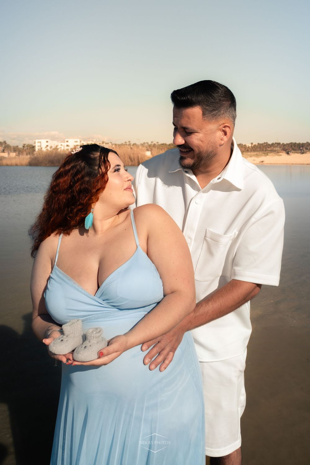 Pregnant couple in light blue and white outfits holding baby shoes by a serene beach at golden hour.