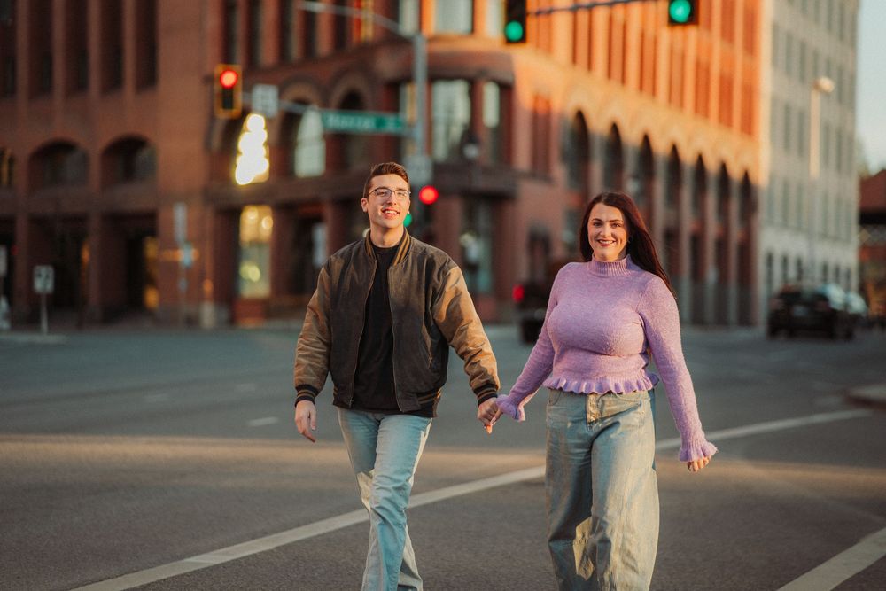 Couple holding hands crossing a city street at sunset, woman in purple turtleneck and man in bomber jacket.