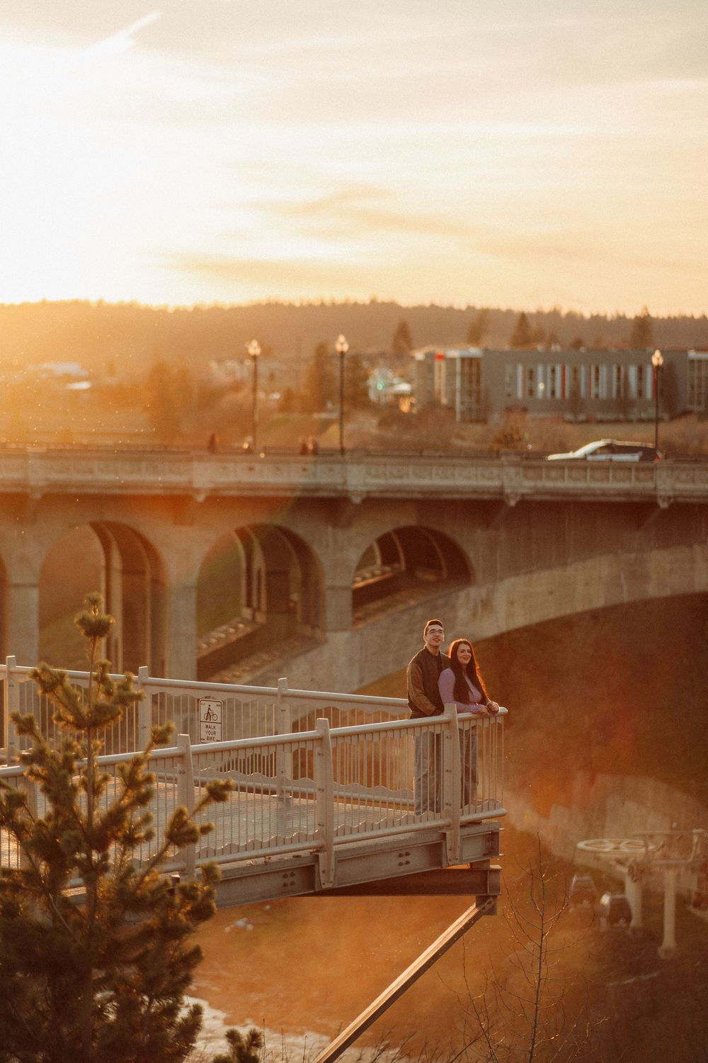 A couple stands on a bridge overlook at golden hour, with a large arched bridge and city skyline behind them.