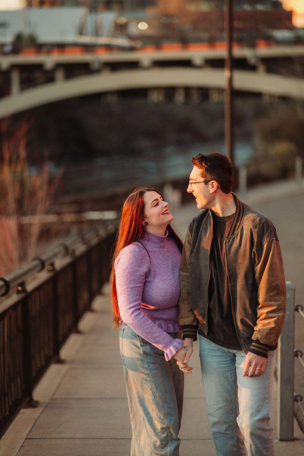 Happy couple holding hands and smiling at each other while walking along a riverside path at sunset.