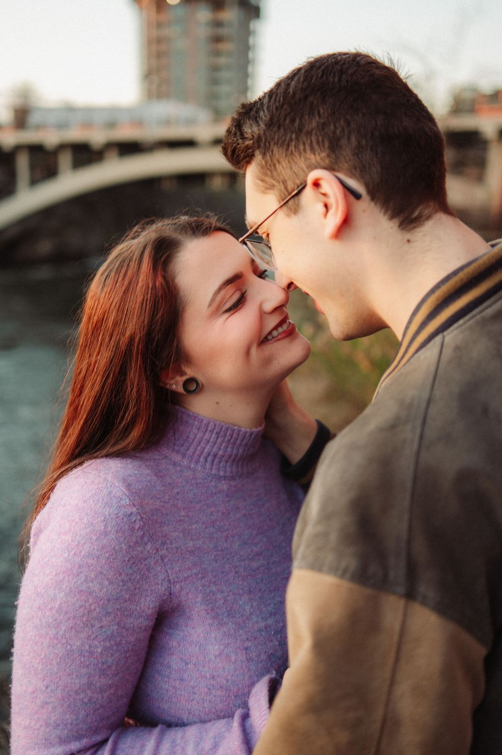 Romantic couple sharing a close moment near a bridge, smiling and nearly kissing at golden hour.