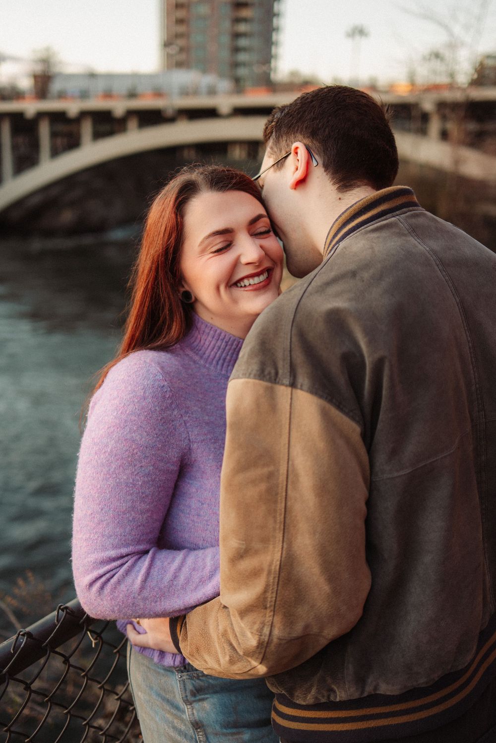 A laughing woman enjoys a tender moment with her partner by a river bridge at sunset.