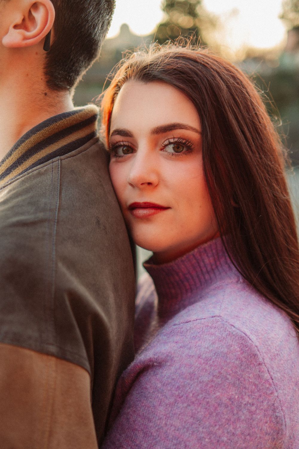 A young woman with long brown hair leans her head on a man's shoulder outdoors in warm golden light.