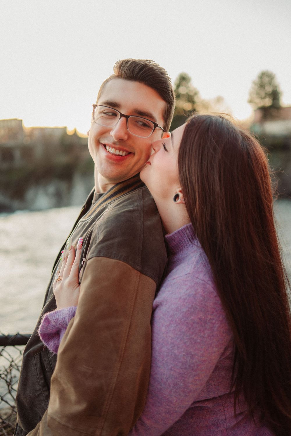 A woman in a pink sweater kisses a smiling man in glasses outdoors near a river at golden hour.