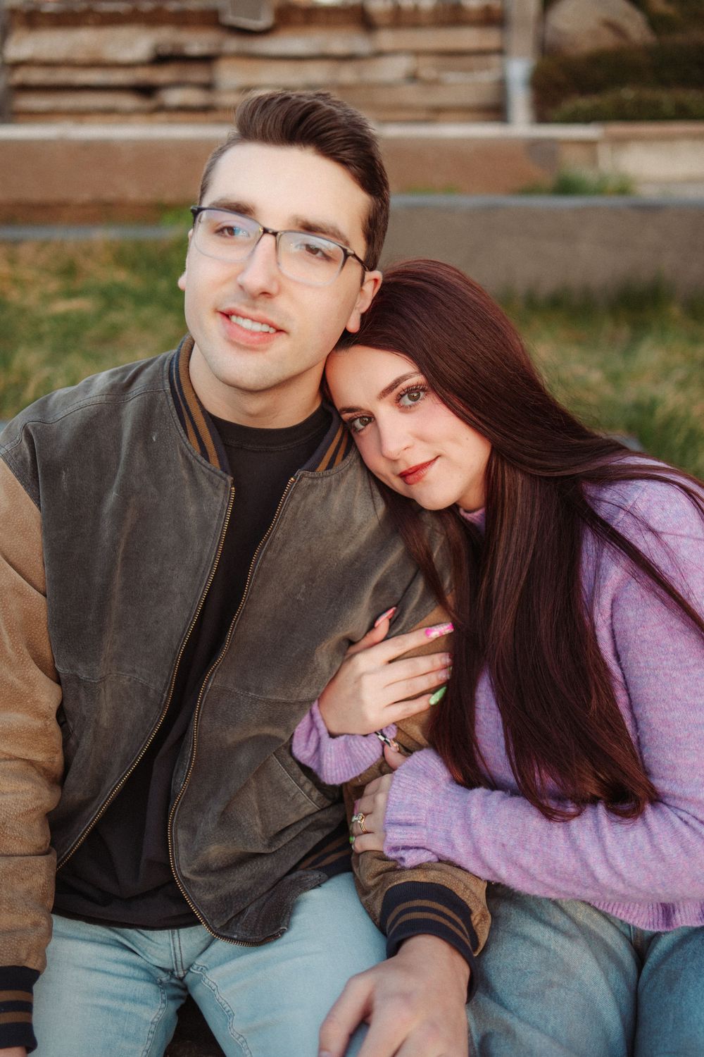 A smiling couple sitting outdoors, woman leaning her head on mans shoulder affectionately.