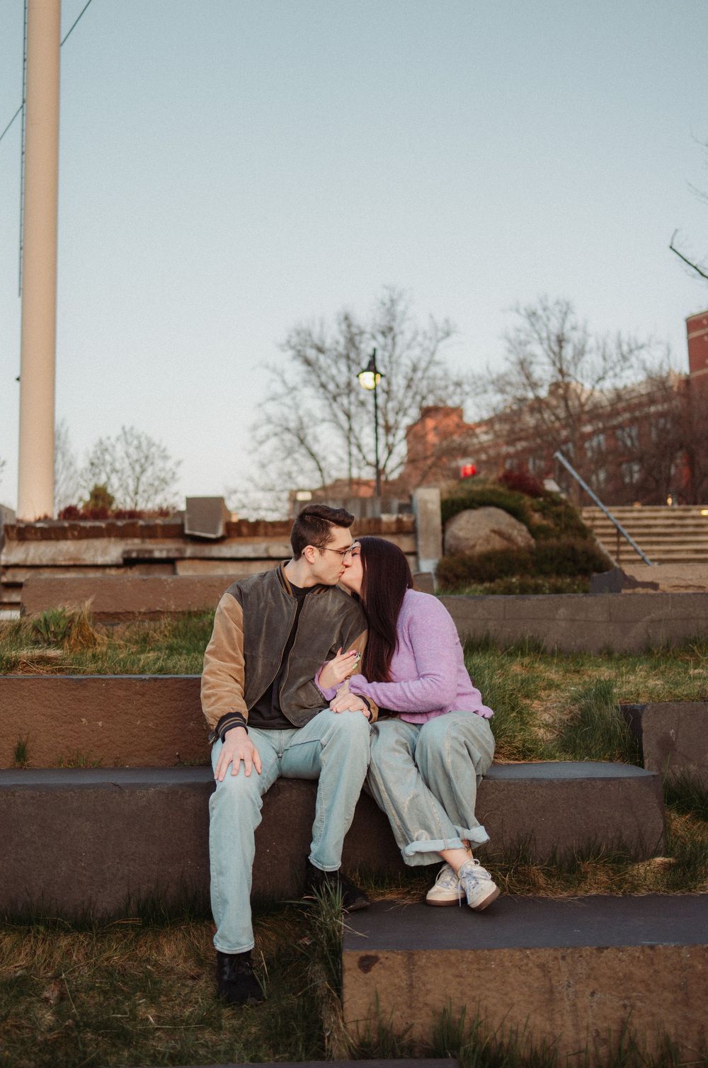 Couple sitting close together on outdoor steps at dusk, sharing a tender kiss in urban park setting.