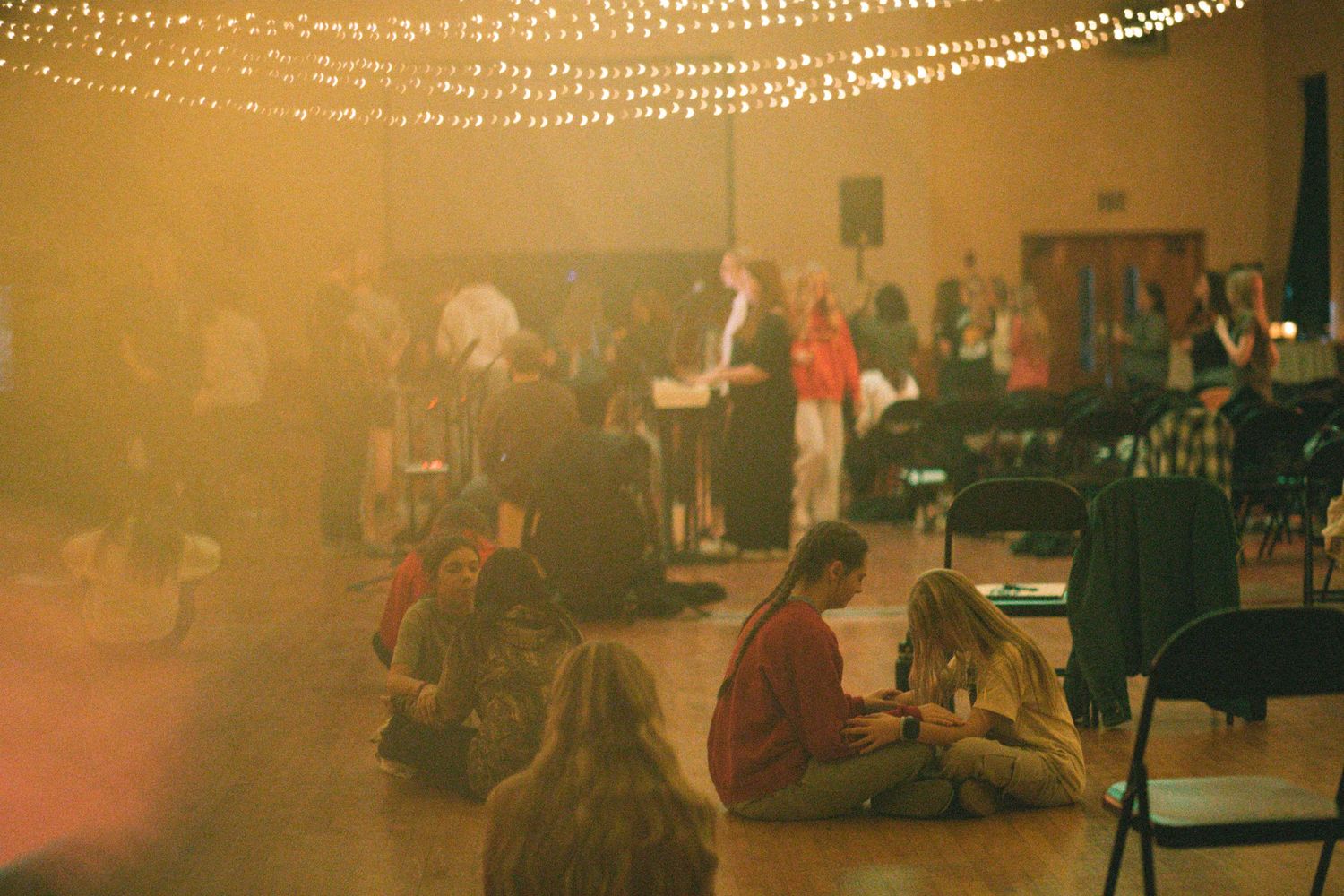 People gathered in warmly lit event hall with string lights and someone presenting at front.