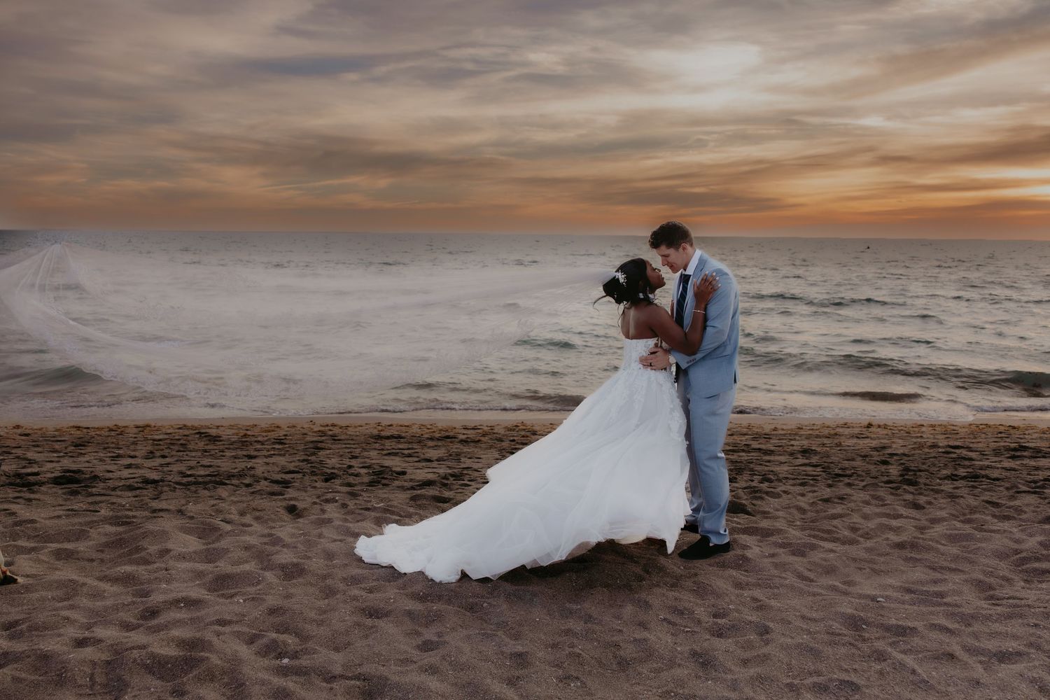 Bride and groom embracing by tall windows in soft natural light, bride wearing lace gown with flowing veil.