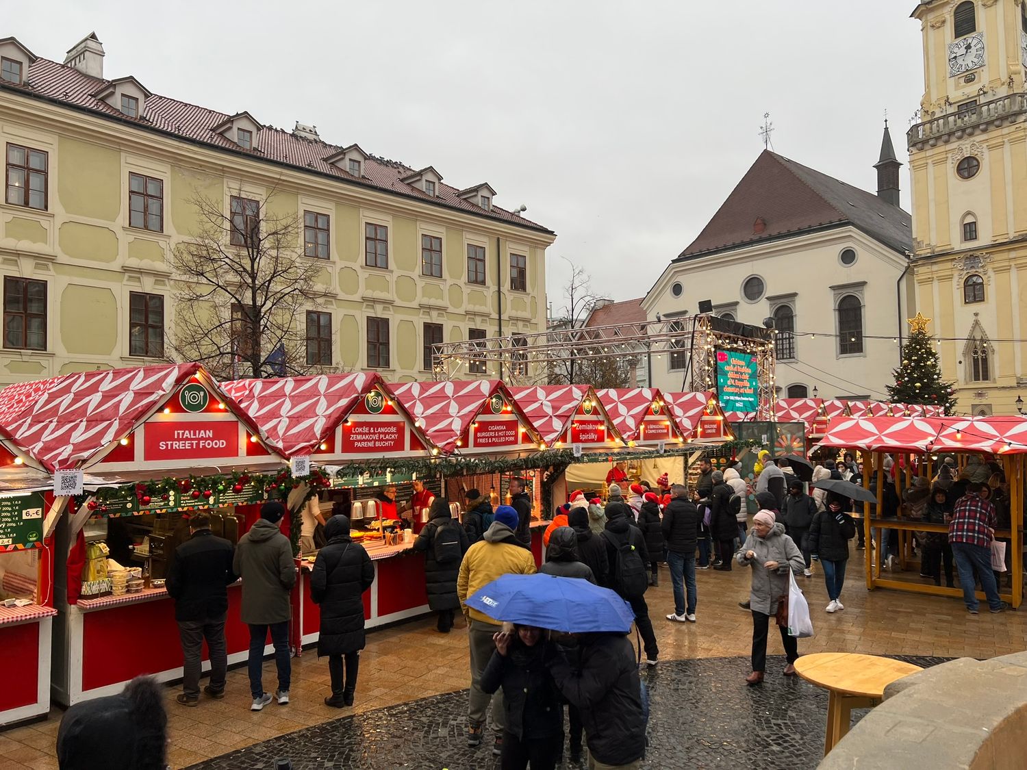Red-roofed Christmas market stalls crowded with shoppers in a historic European town square on a rainy winter day.