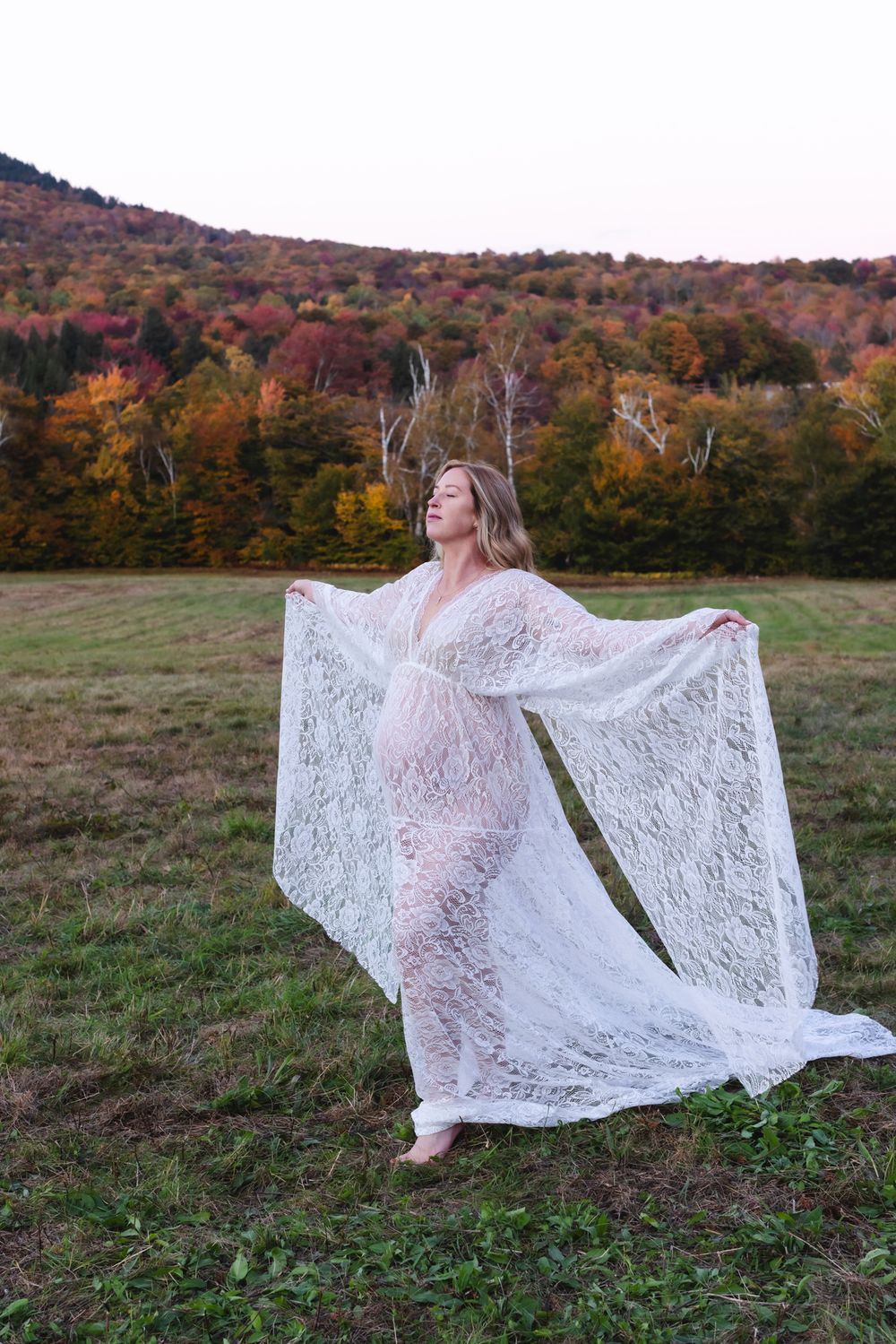 Pregnant mother wears white lace maternity dress walking in open field holding up dress against mountain background. North Conway maternity