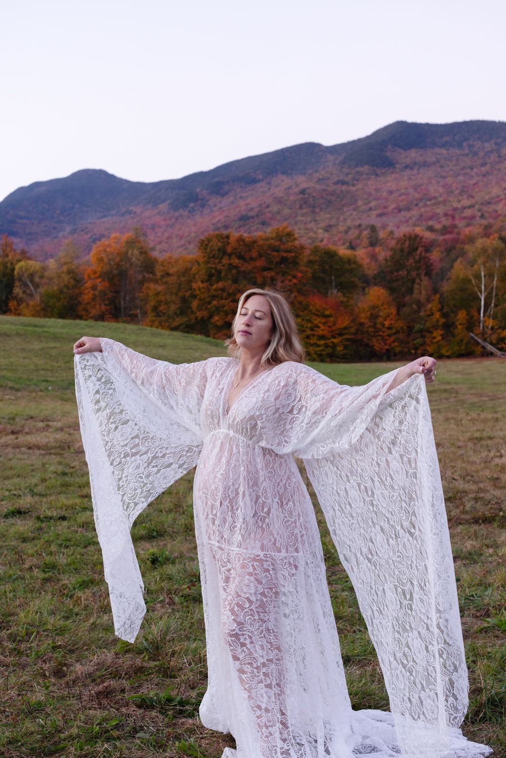 Pregnant mother wears white lace dress posing in open field with mountains and fall foliage background for maternity photos in Stowe, VT.