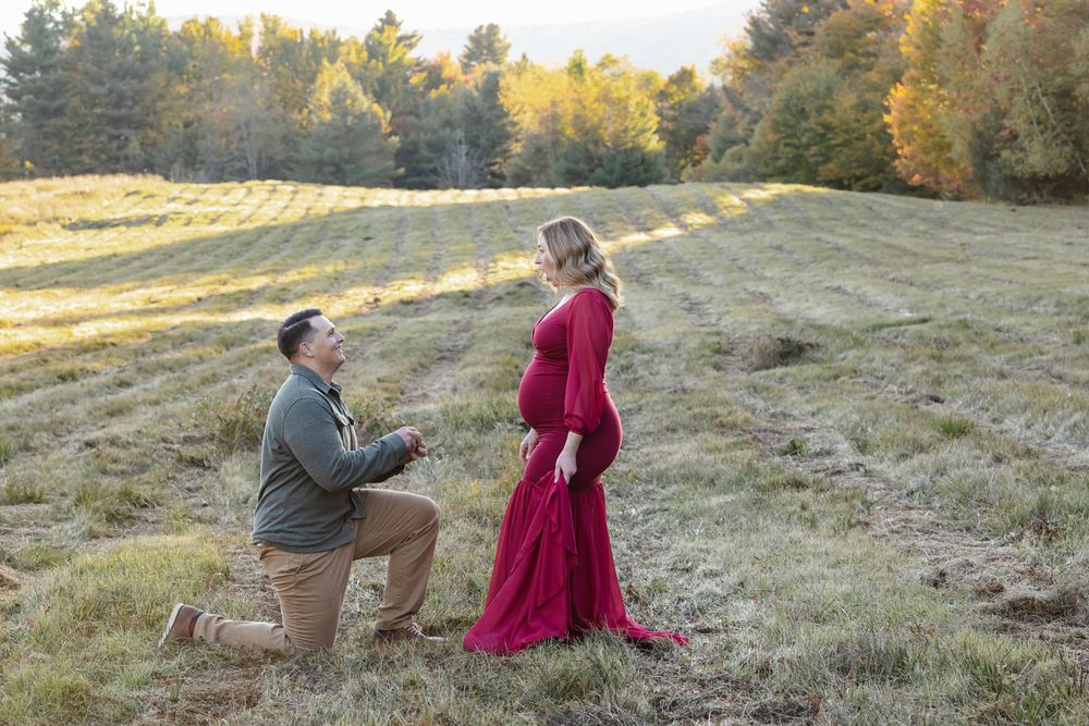 Expectant mother wears a red maternity gown as man proposes to her in an open golden field. North Conway, Lincoln maternity photographer