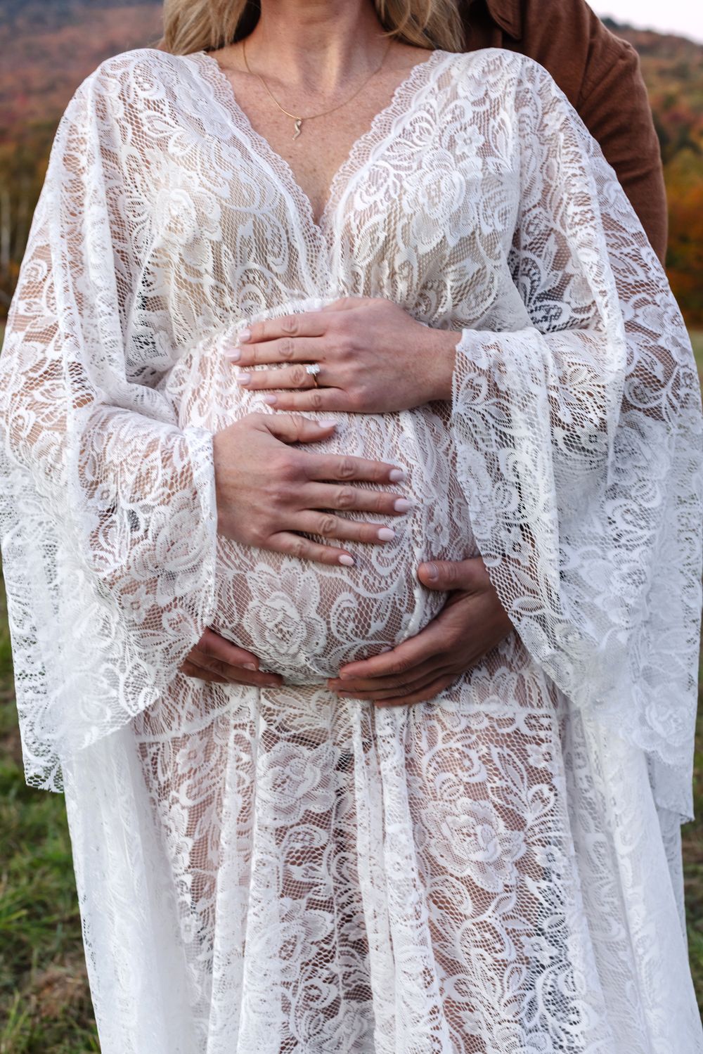 Expectant mother wears white lace maternity gown as fiancee embraces from behind for a close up of baby bump. North Conway maternity photos
