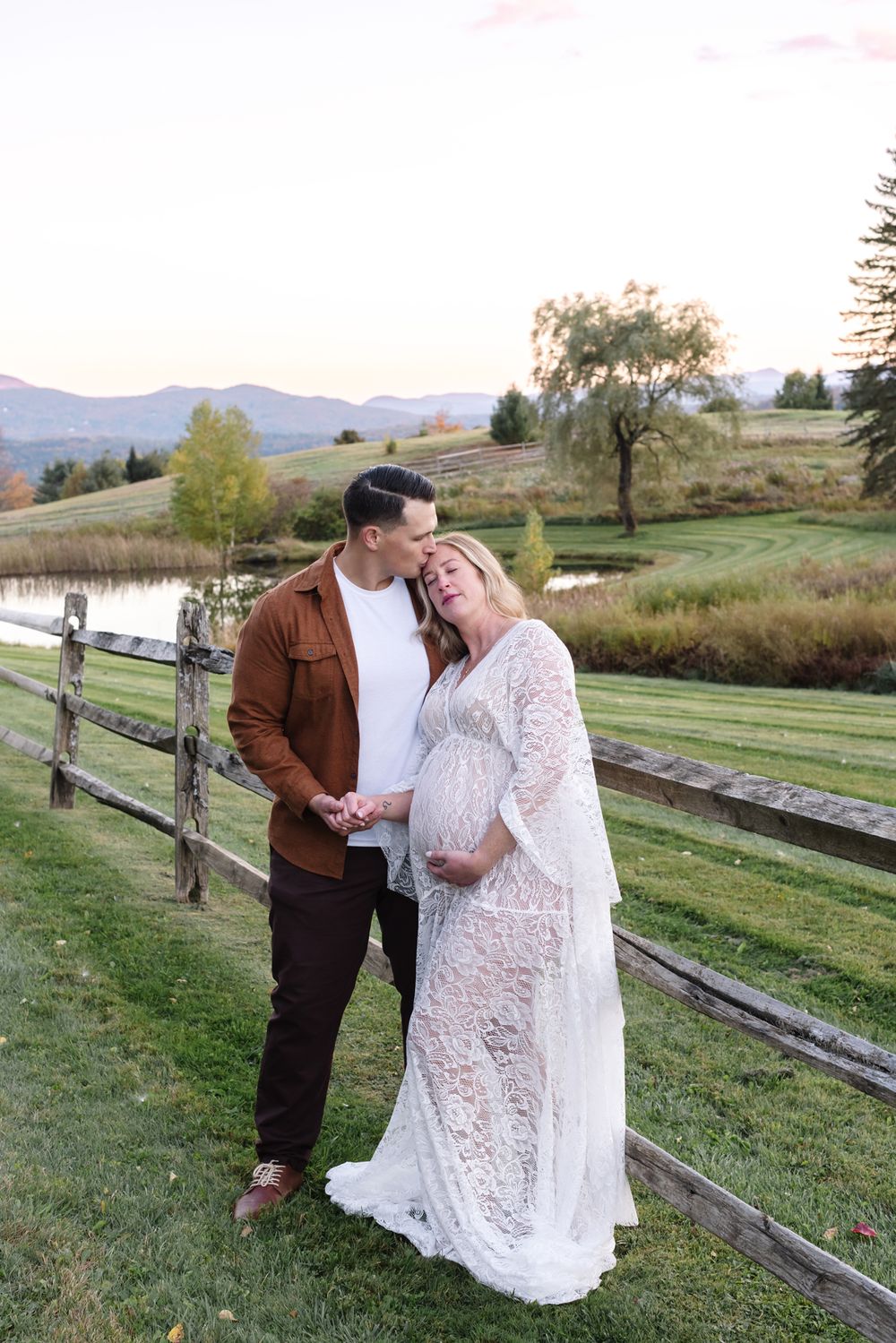 Expectant mother kissed on forehead by fiancee posing by wooden fence and mountains in background. Berkshires maternity photographer