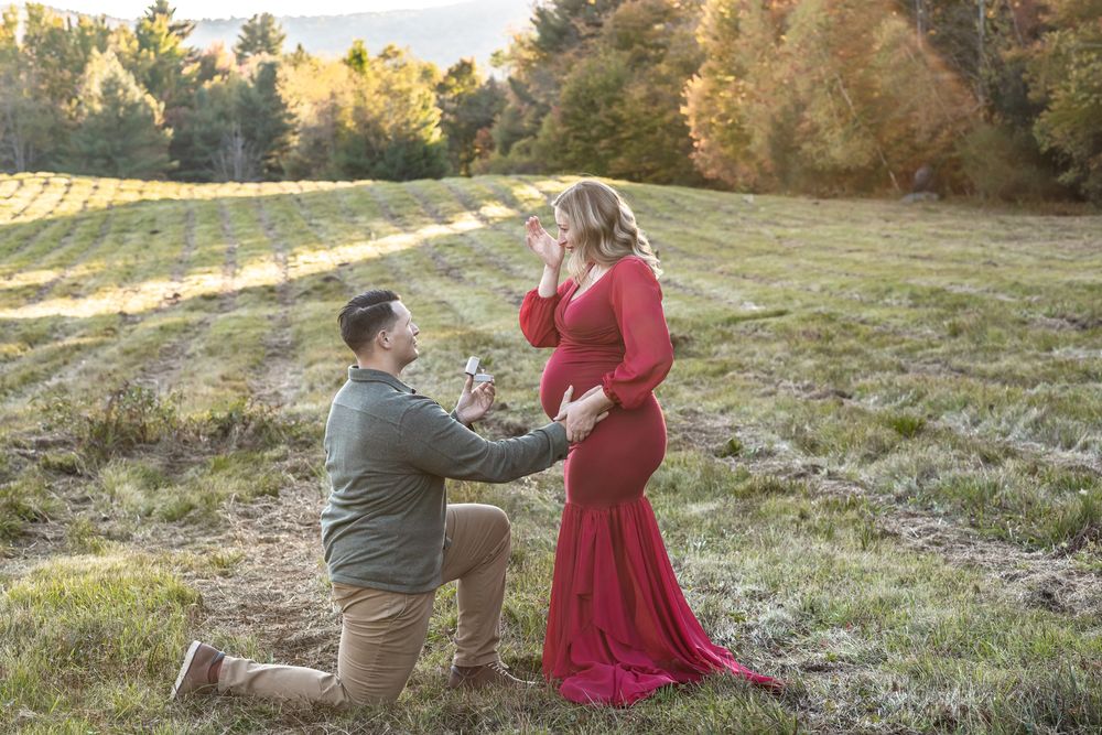 Expectant mother in red maternity gown is propped to in an open field with golden light in Stowe, Vermont for maternity engagement shoot.