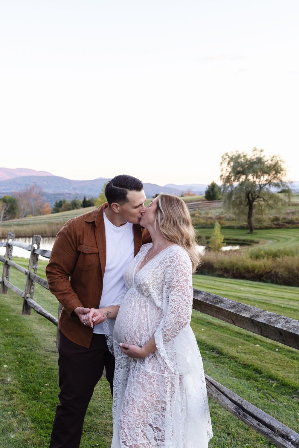 Expectant mother in white lace gown kisses fiancee next to wooden fence with mountains in background for maternity photos in Stowe, VT.
