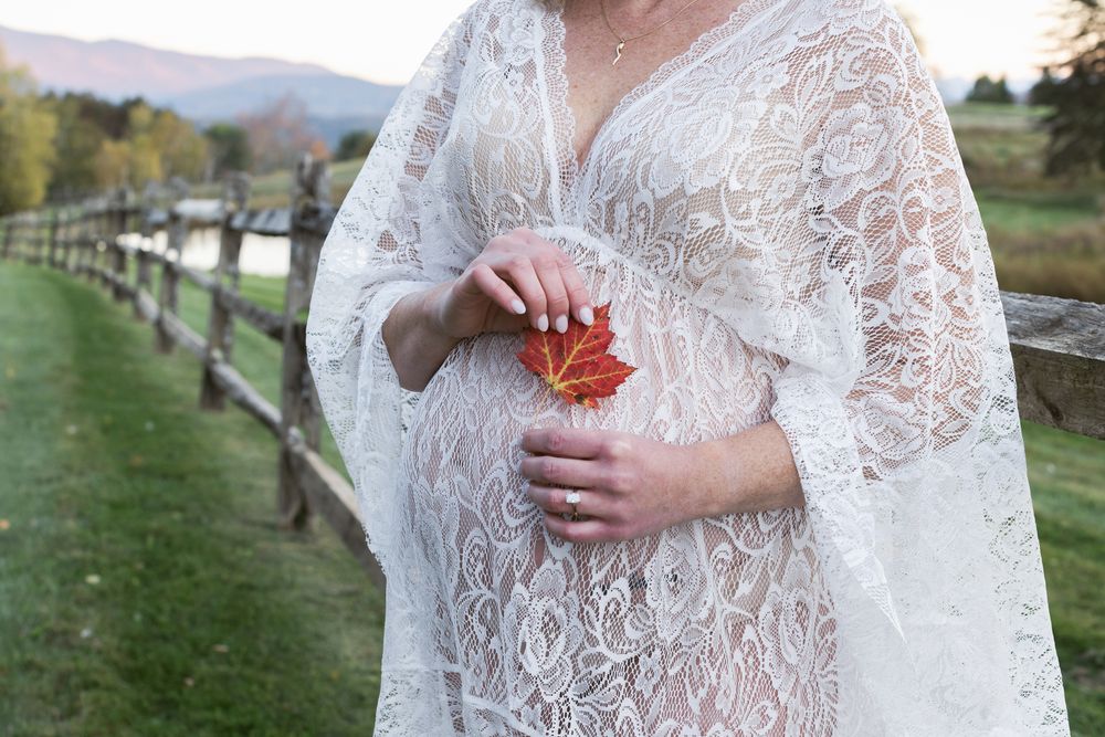 Expectant mother wears white lace dress holding a red leaf against baby bump in front of wooden fence. North Conway maternity photographer