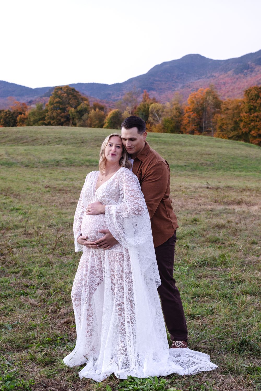 Expectant mother wears white lace dress with fiancee embracing from behind her in an open field with mountain and fall foliage backdrop.