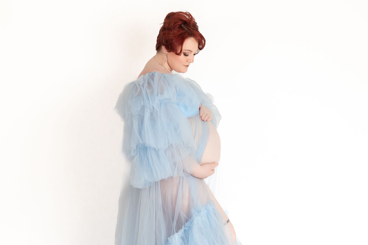 Pregnant woman in sheer blue ruffled maternity gown cradling baby bump against white background.
