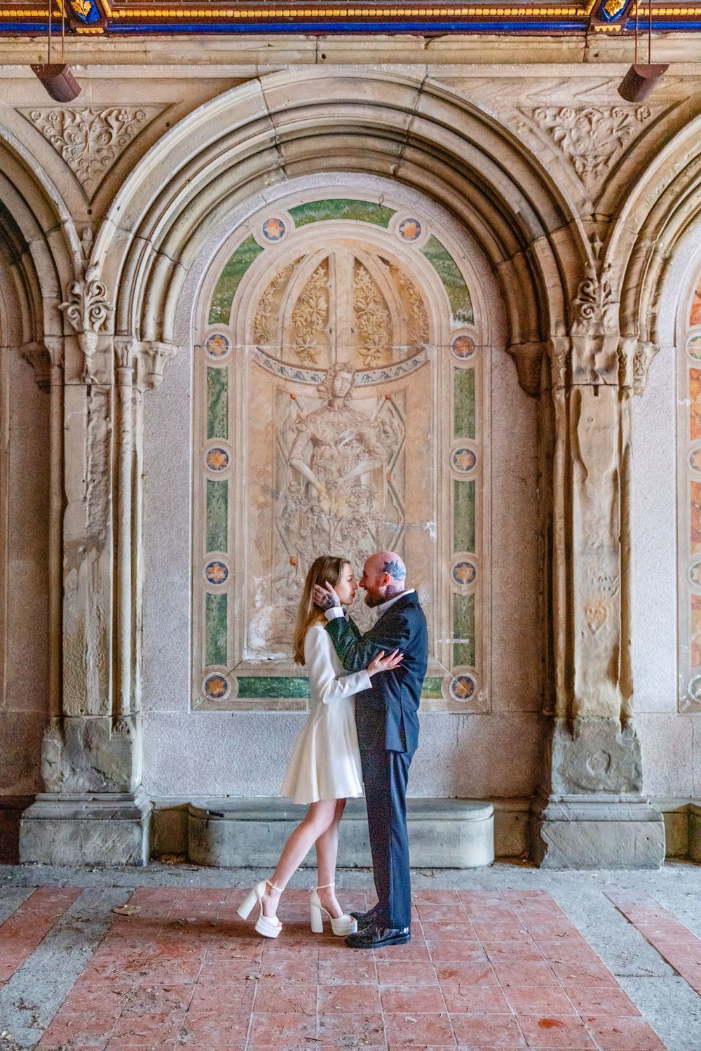 Couple embracing under ornate stone arches with mosaic mural at Bethesda Terrace, Central Park, NYC.