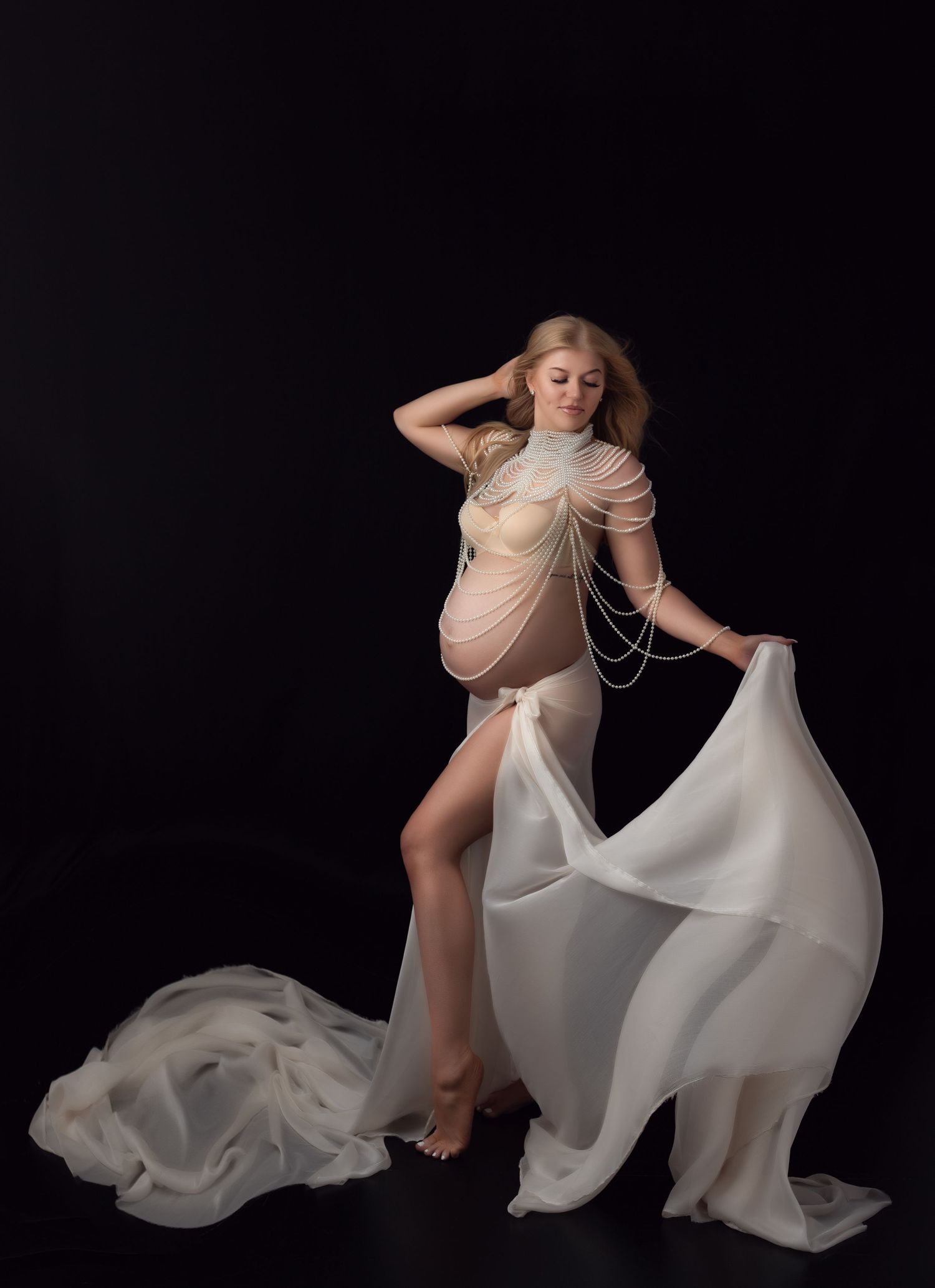 A family luxury style maternity photoshoot features a couple in beige outfits with a child on a stool against a dark backdrop.