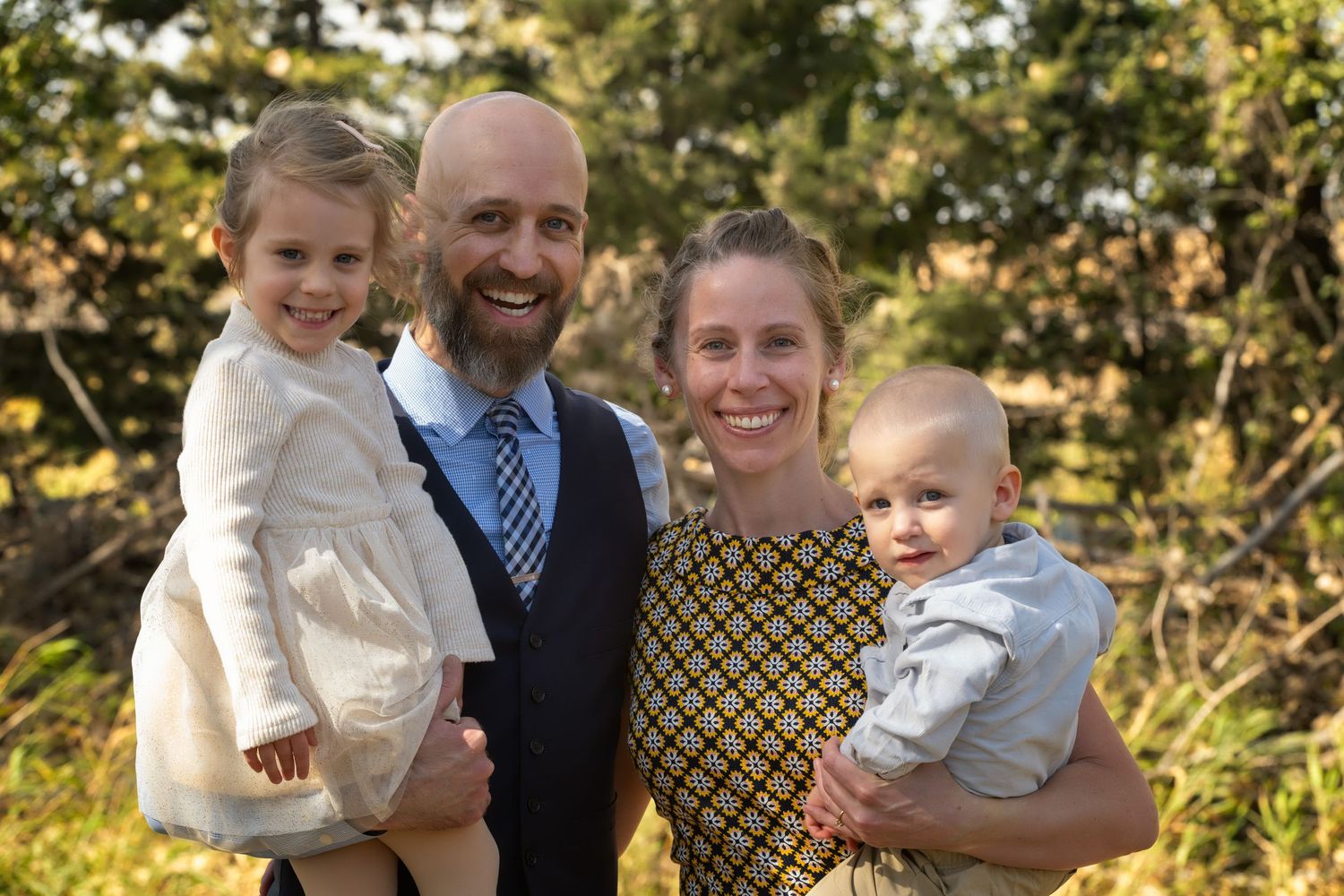 Happy family of four posing outdoors in autumn, parents holding two young children, smiling in a wooded setting.