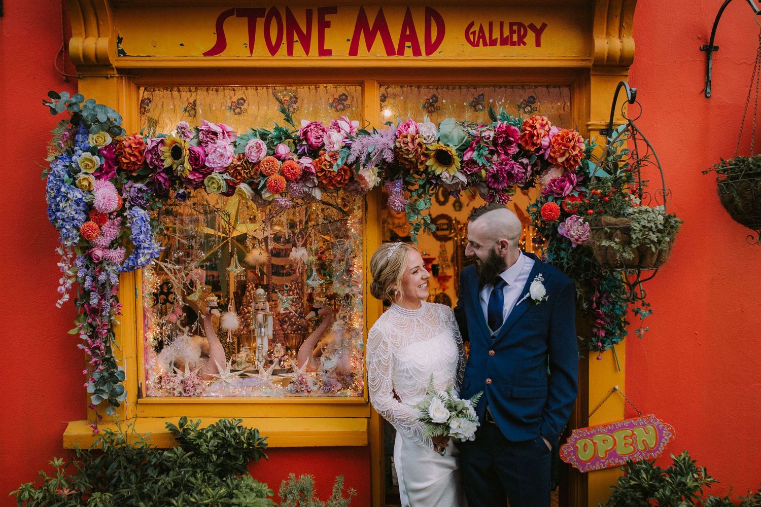 Bride and groom pose in front of colorful Stone Mad Gallery adorned with vibrant flowers in Ireland.